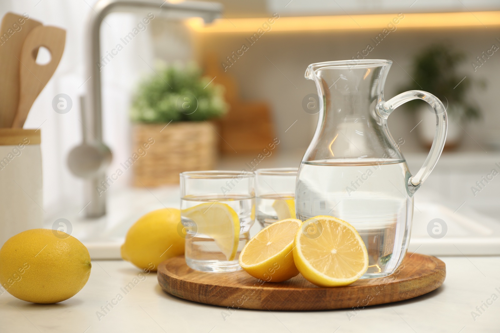 Photo of Jug, glasses with clear water and lemons on white table in kitchen