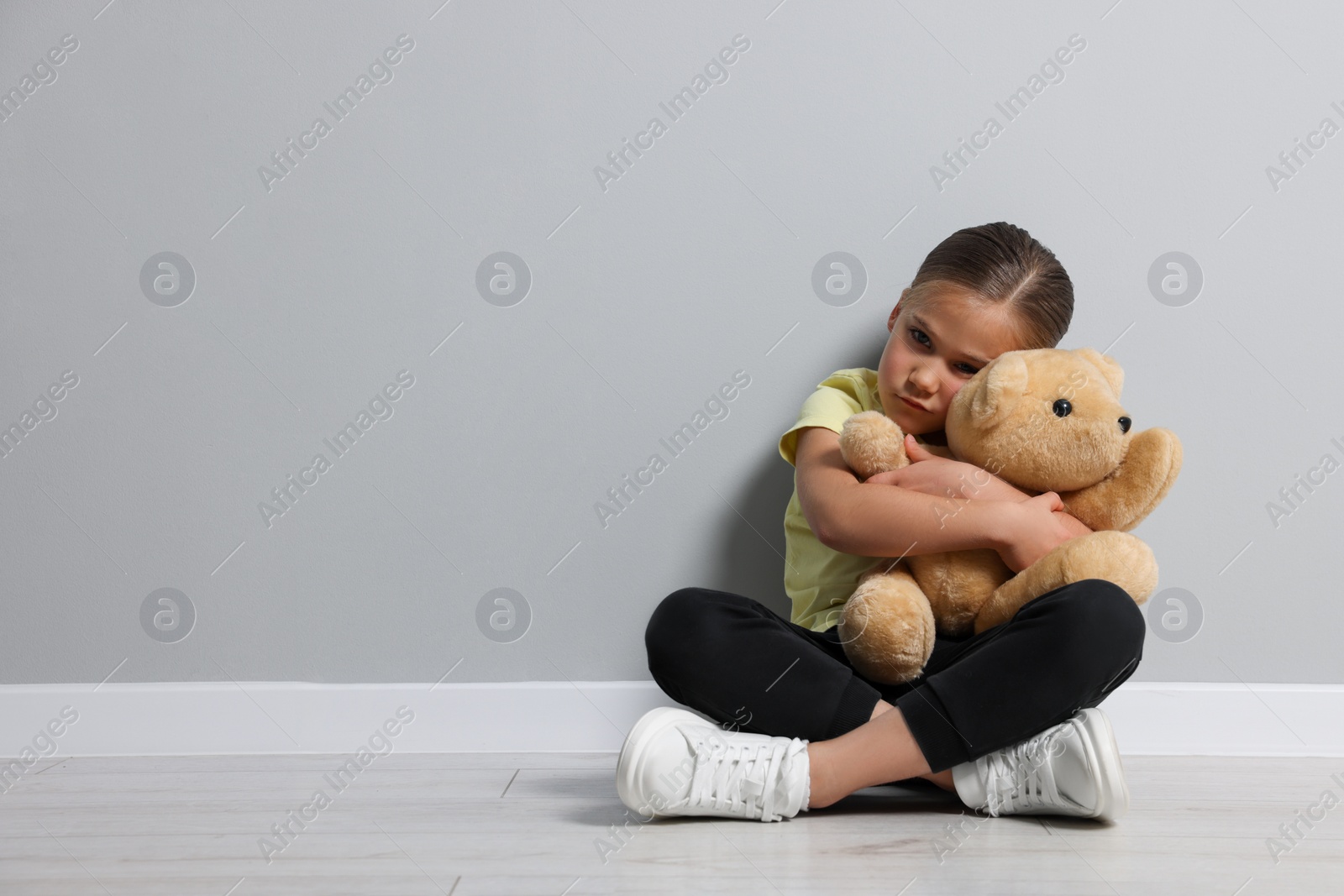 Photo of Child abuse. Upset girl with toy sitting on floor near grey wall, space for text