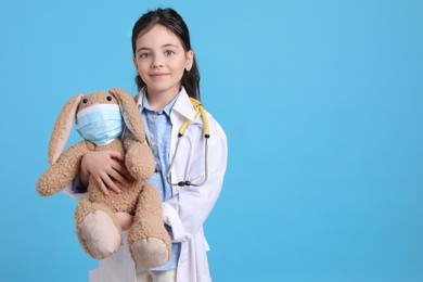 Photo of Little girl with stethoscope and toy bunny dressed as doctor on light blue background, space for text. Pediatrician practice