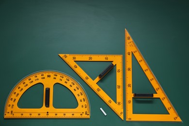 Photo of Protractor, triangles and chalk on green chalkboard, flat lay