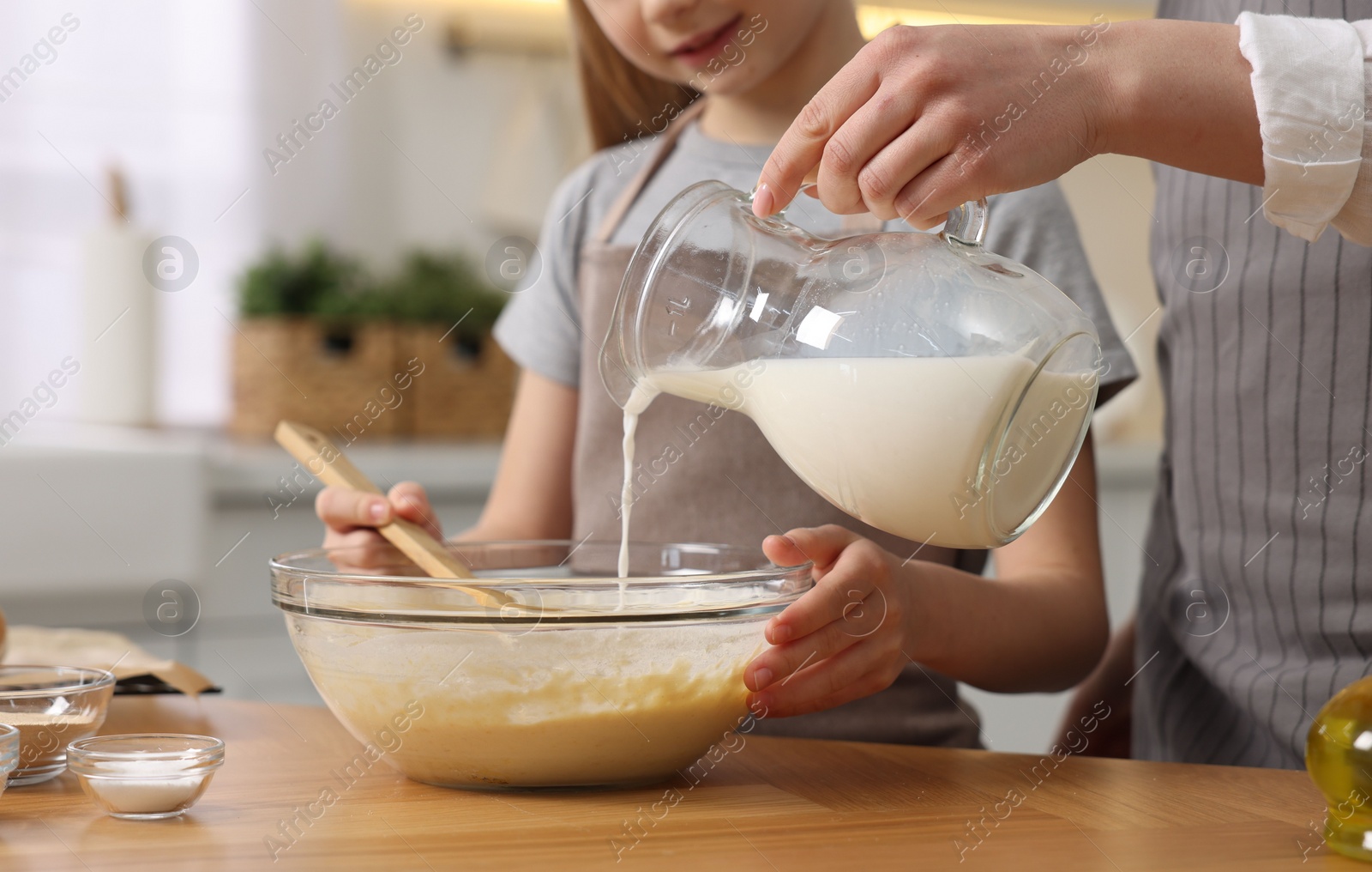 Photo of Making bread. Mother and her daughter pouring milk into bowl at wooden table in kitchen, closeup