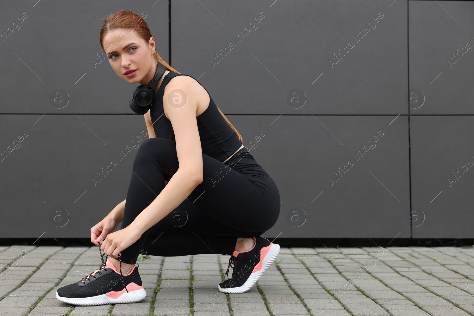 Photo of Beautiful woman in stylish gym clothes with headphones tying shoelace of sneakers near dark grey wall on street, space for text