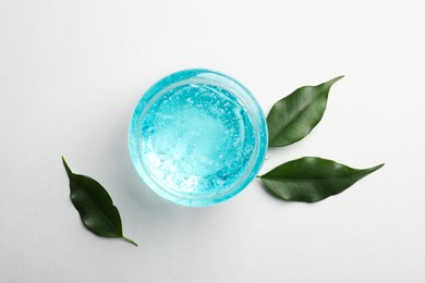 Photo of Jar of blue cosmetic gel and green leaves on white background, flat lay