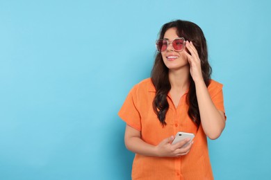 Happy beautiful woman with stylish sunglasses and smartphone on light blue background, space for text