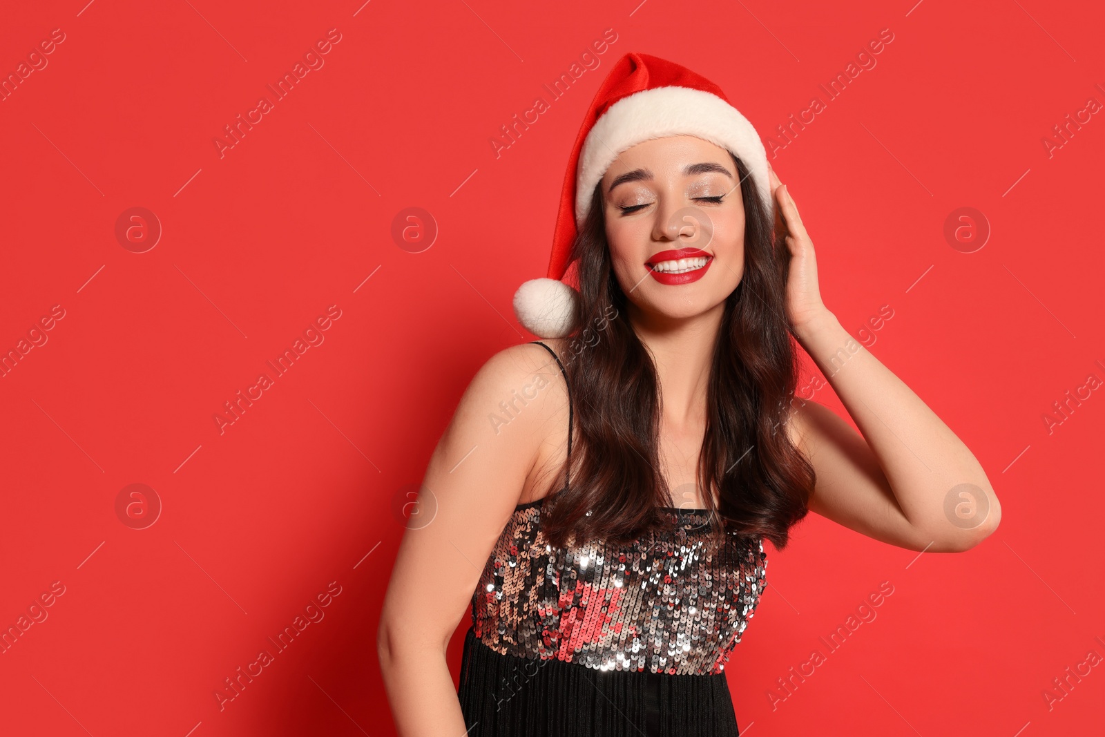 Photo of Christmas celebration. Beautiful young woman in stylish dress and Santa hat on red background, space for text