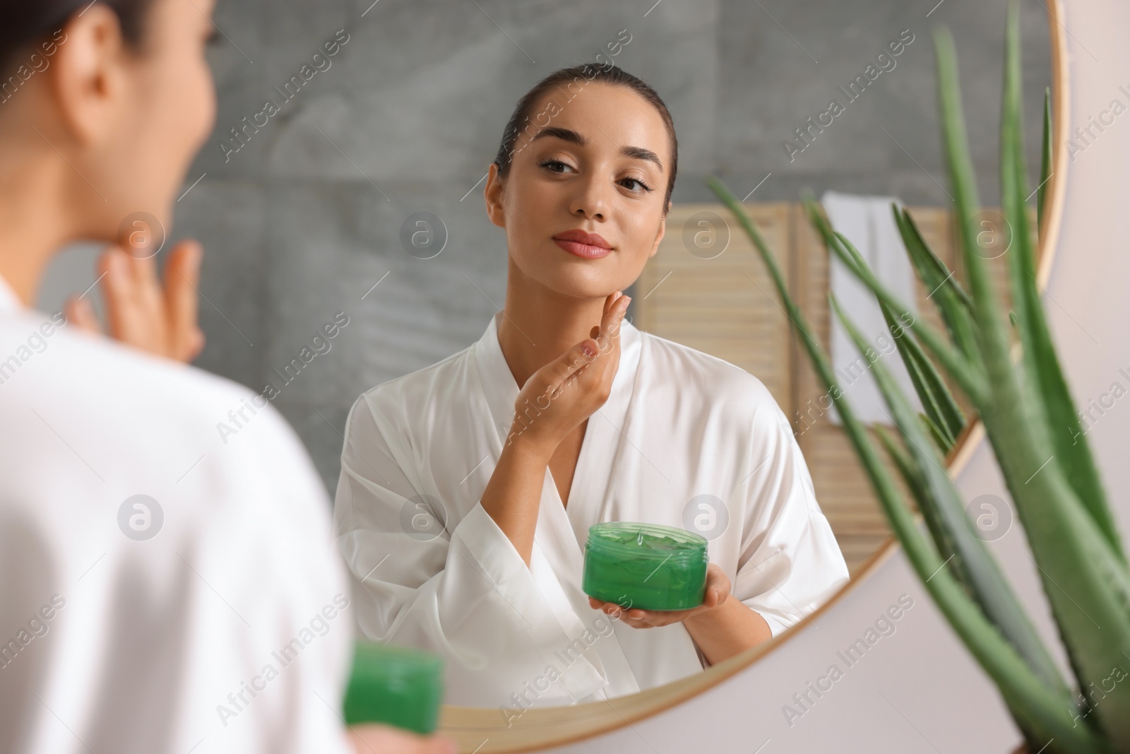 Photo of Young woman holding jar of aloe gel near mirror in bathroom