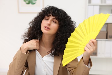 Young businesswoman waving yellow hand fan to cool herself in office