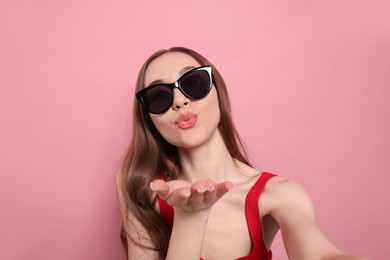 Beautiful young woman with sunglasses blowing kiss while taking selfie on pink background