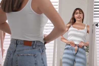 Photo of Young woman trying to put on tight jeans near mirror at home