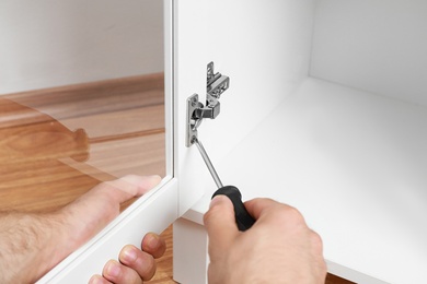 Photo of Man fixing door angle hinge of wooden cabinet, closeup view