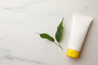 Face cream in tube and leaves on white marble table, flat lay. Space for text