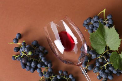 Photo of Overturned glass with red wine and grapes on brown background, flat lay