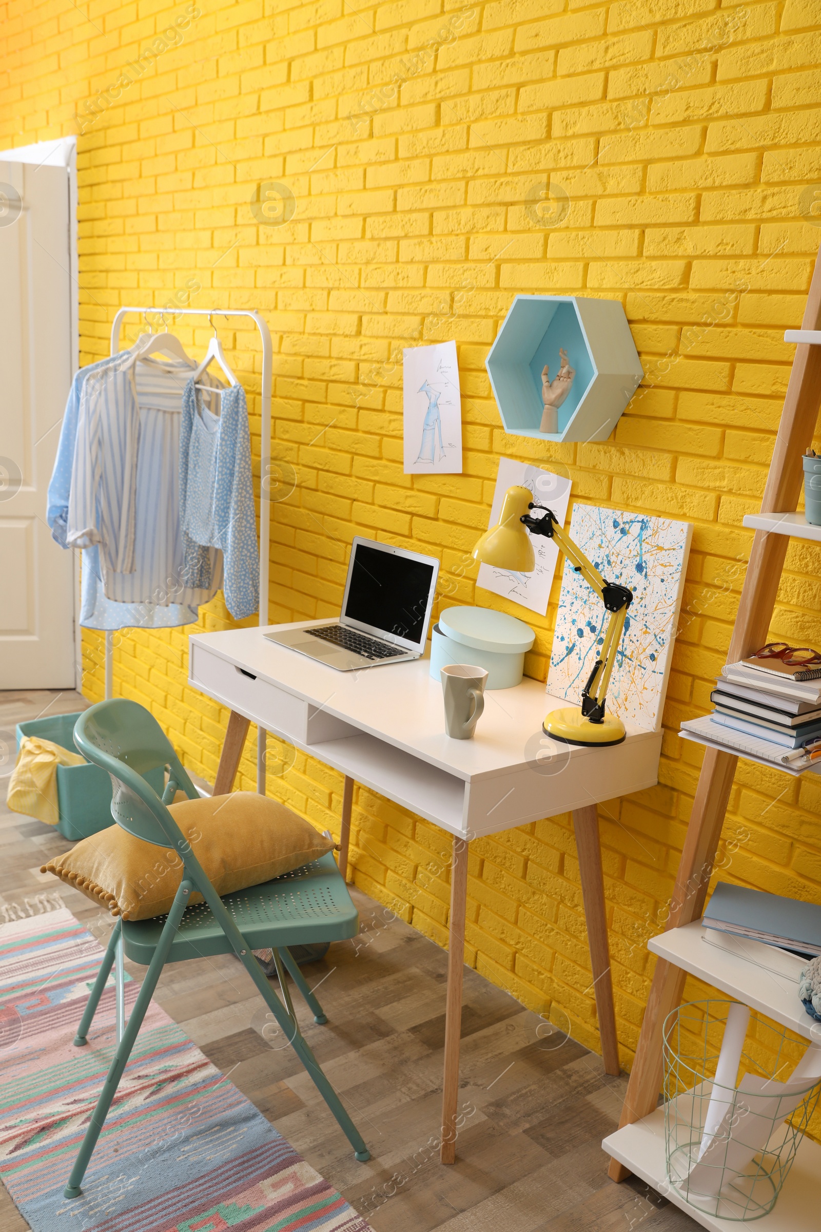 Photo of Fashion designer's workplace with wooden furniture and laptop near yellow brick wall. Stylish interior