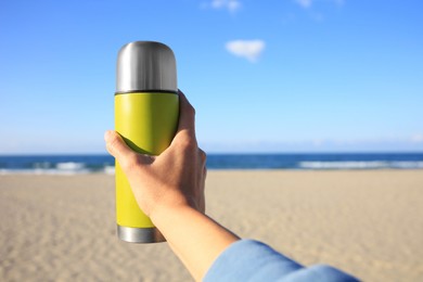 Photo of Woman holding yellow thermos with hot drink on beach near sea, closeup. Space for text