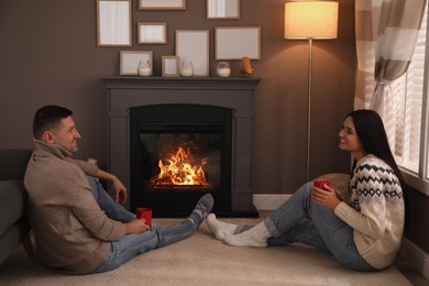 Photo of Lovely couple with cups of hot drink spending time together near fireplace at home
