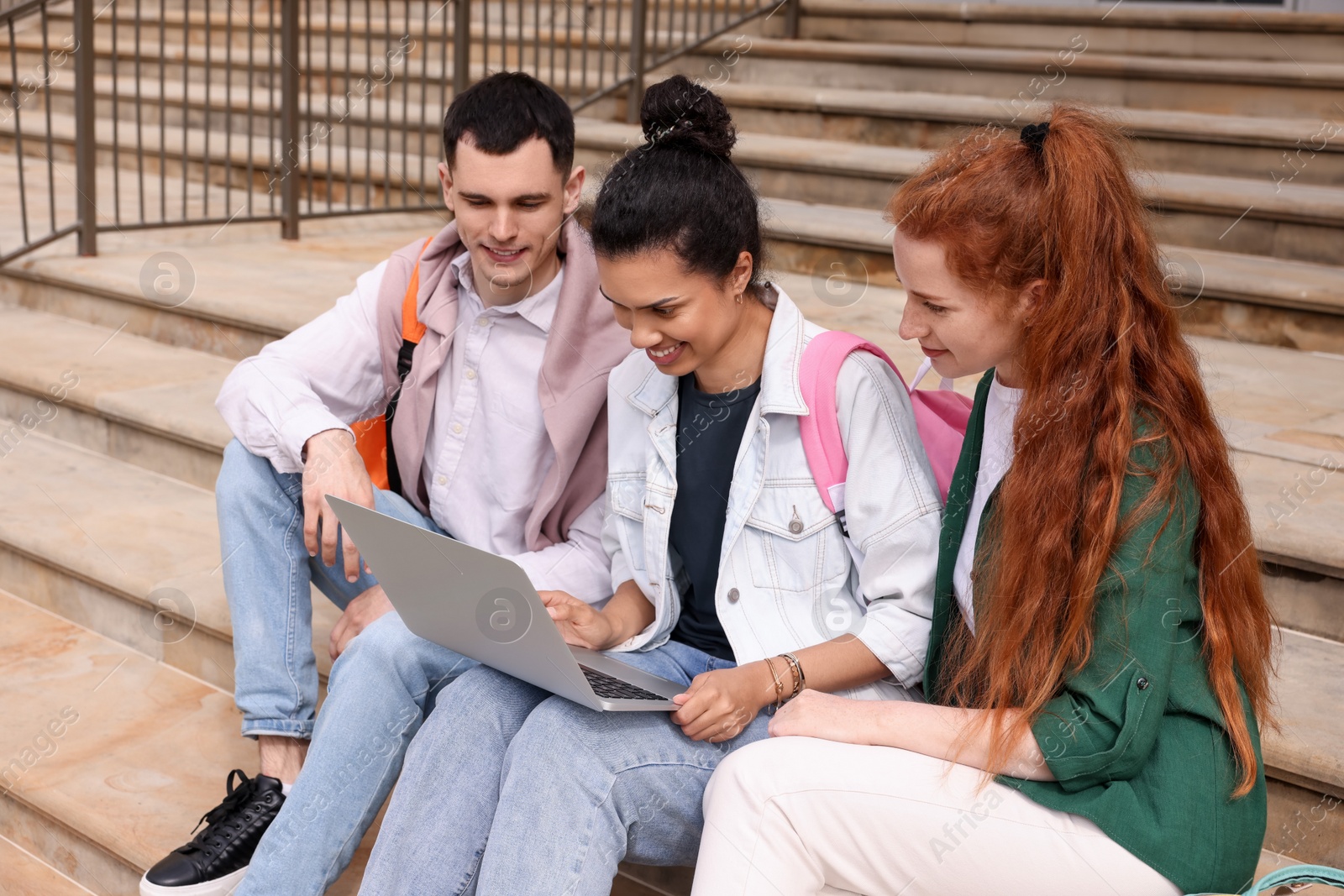 Photo of Happy young students studying together with laptop on steps outdoors