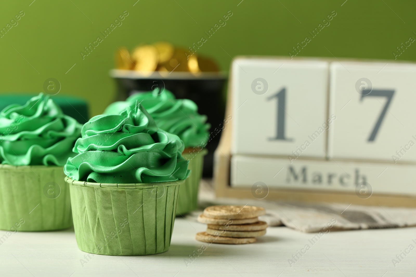 Photo of St. Patrick's day party. Tasty cupcakes with green cream, pot of gold and wooden block calendar on white table, closeup