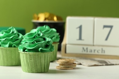 Photo of St. Patrick's day party. Tasty cupcakes with green cream, pot of gold and wooden block calendar on white table, closeup