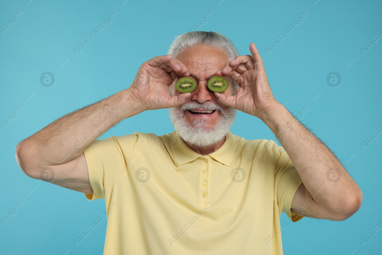 Photo of Senior man covering eyes with halves of kiwi on light blue background