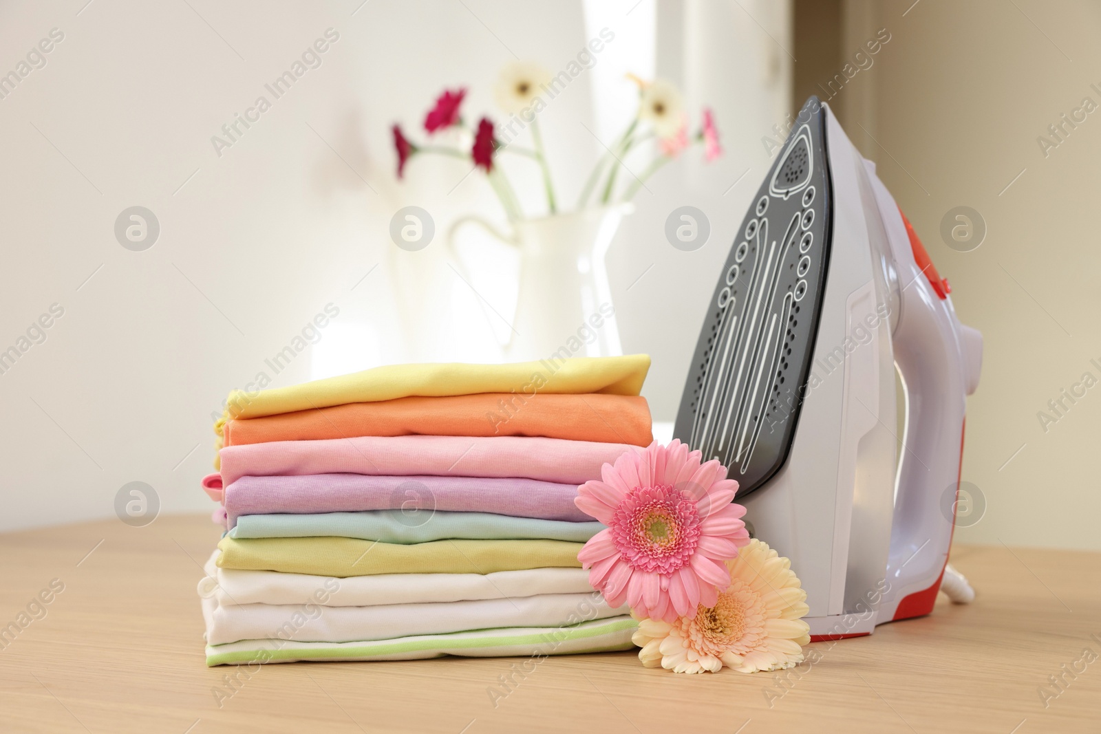 Photo of Stack of clean clothes, modern iron and flowers on wooden table