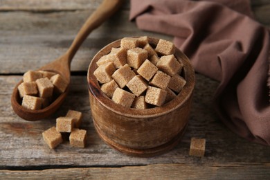 Photo of Brown sugar cubes in bowl and spoon on wooden table, closeup