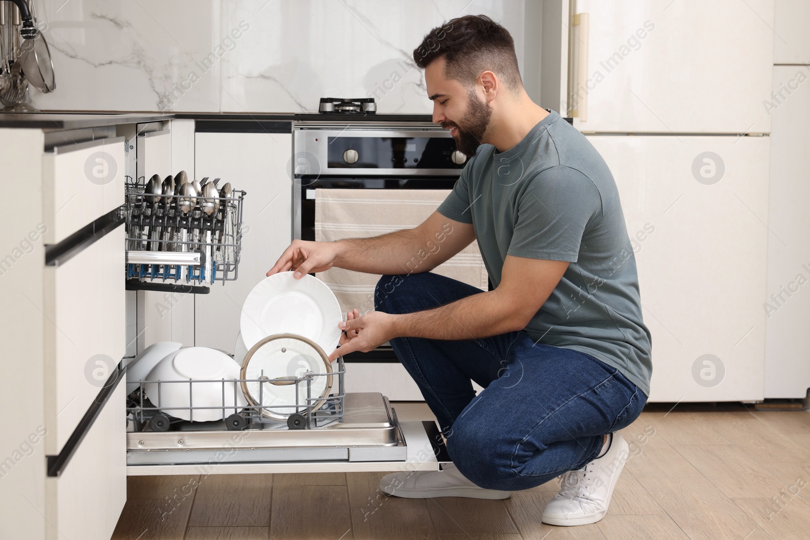 Photo of Smiling man loading dishwasher with plates in kitchen