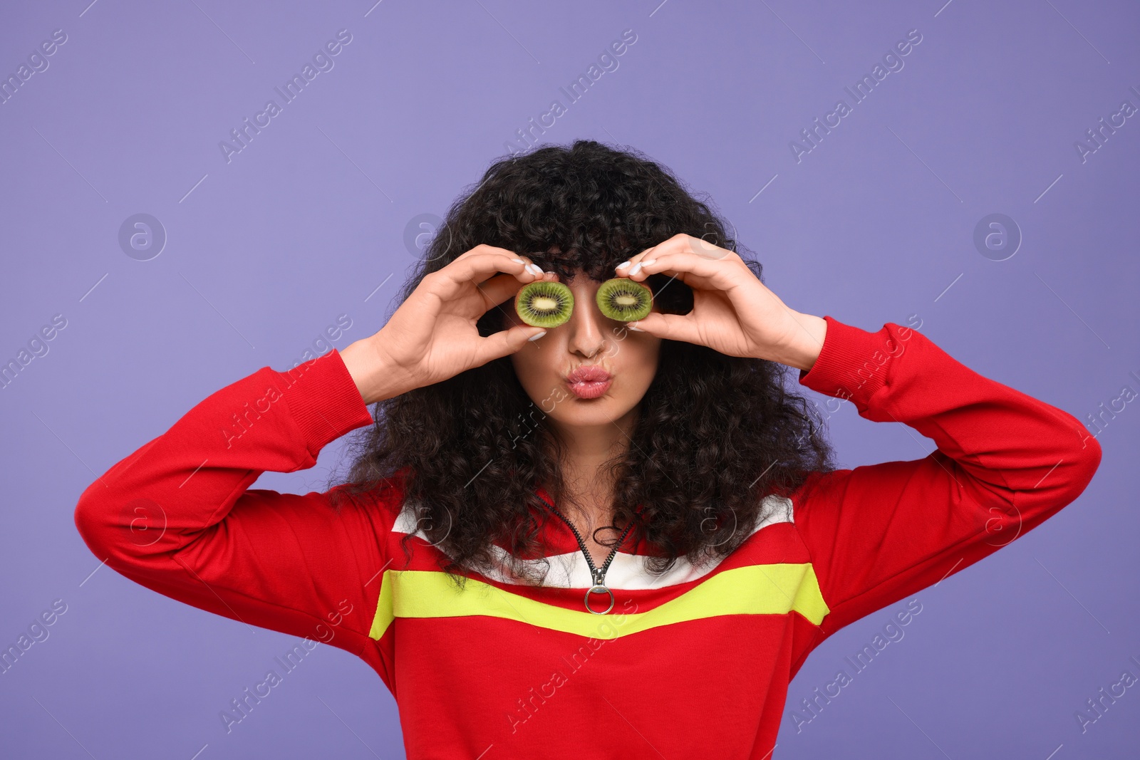 Photo of Woman covering eyes with halves of kiwi and blowing kiss on violet background