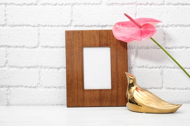 Photo of Blank frame, decorative bird figure and flower on table near brick wall. Mock up for design