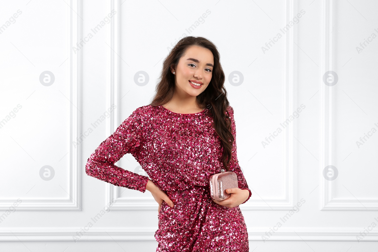 Photo of Beautiful young woman with clutch in stylish pink sequin dress near white wall indoors. Party outfit