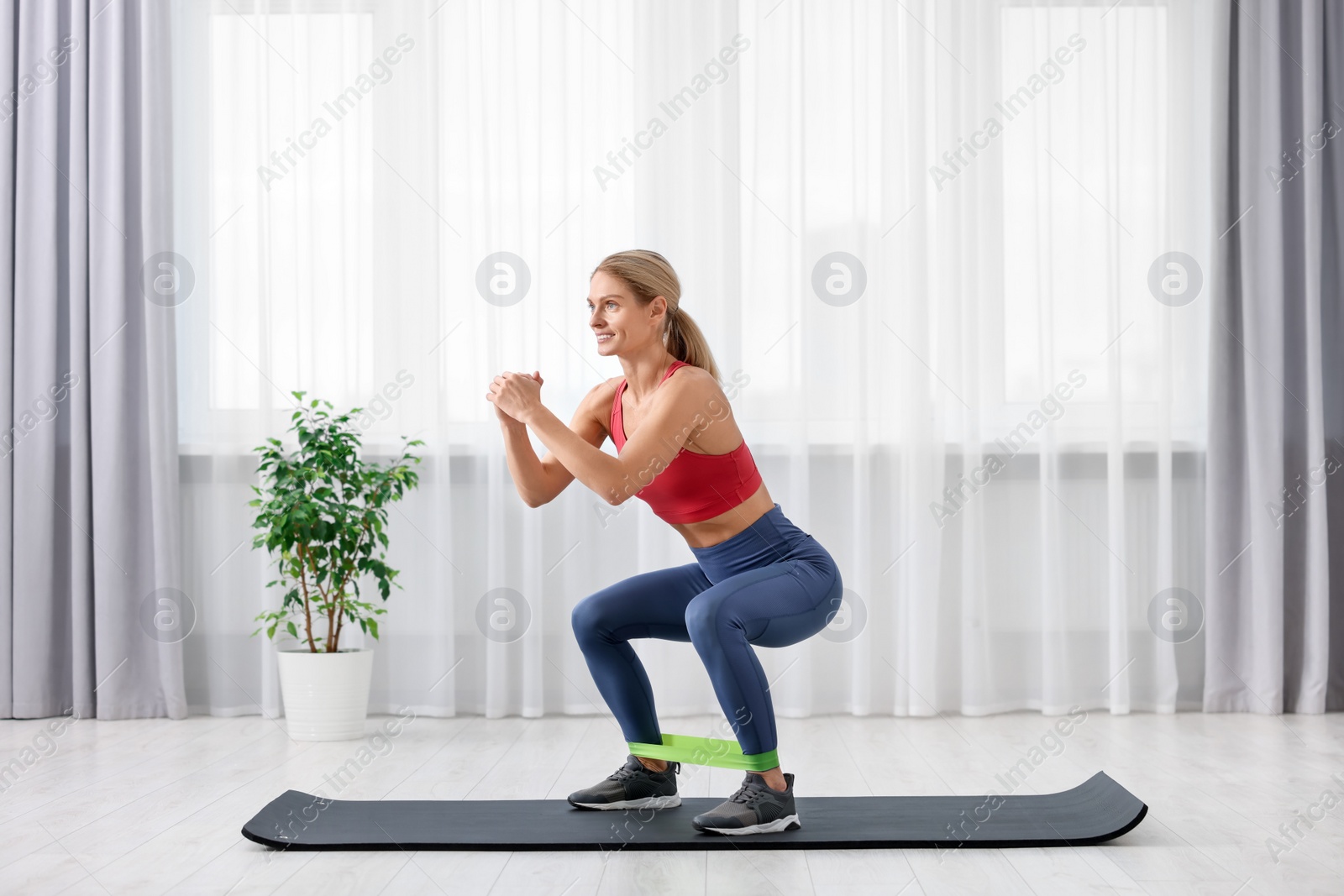 Photo of Athletic woman doing squats with fitness elastic band on mat at home