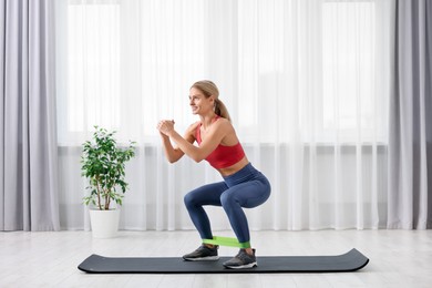 Photo of Athletic woman doing squats with fitness elastic band on mat at home