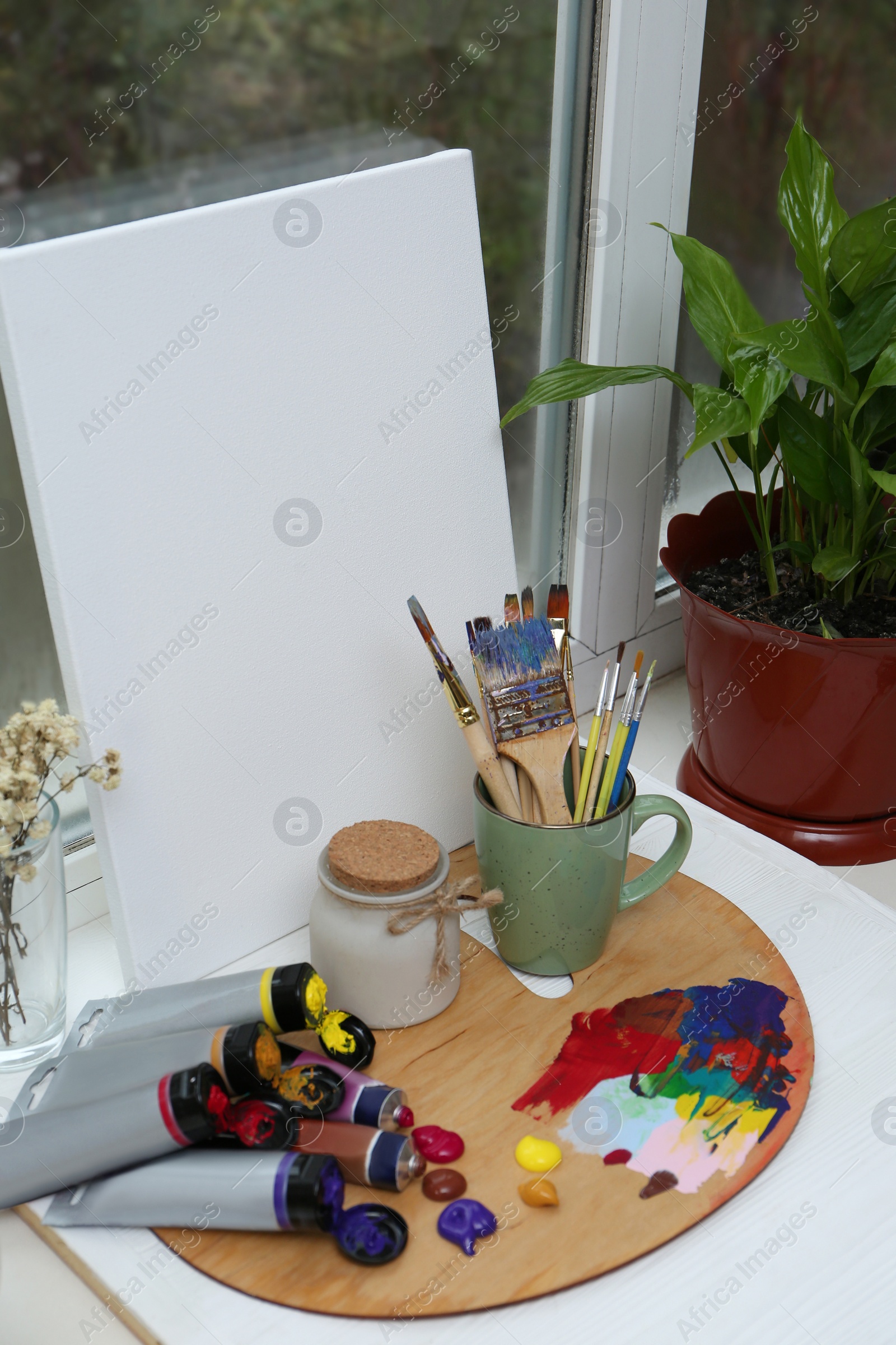 Photo of Wooden artist's palette with colorful paints, brushes and canvas on white windowsill