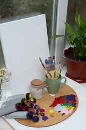 Photo of Wooden artist's palette with colorful paints, brushes and canvas on white windowsill