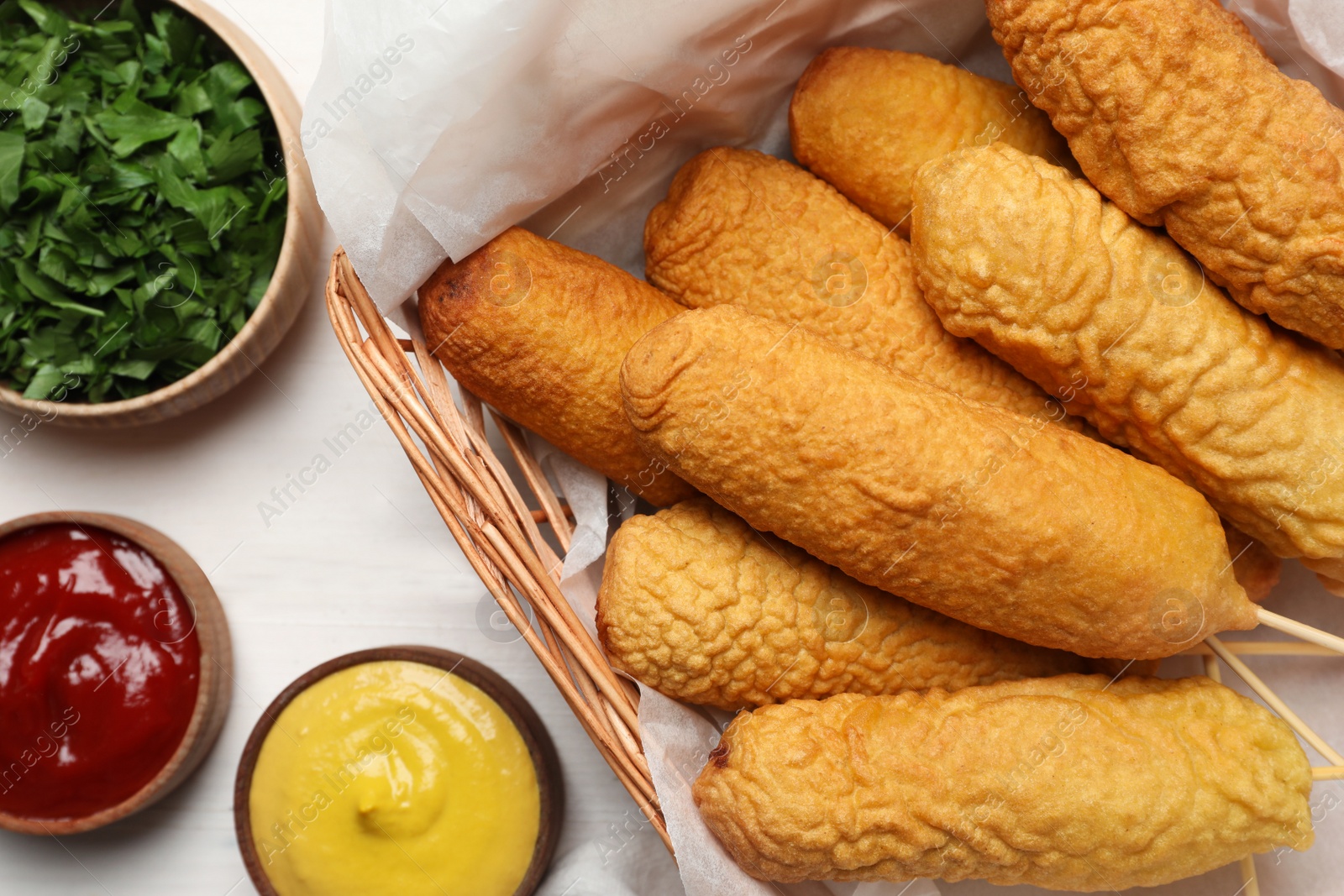 Photo of Delicious deep fried corn dogs in basket and sauces on white wooden table, flat lay