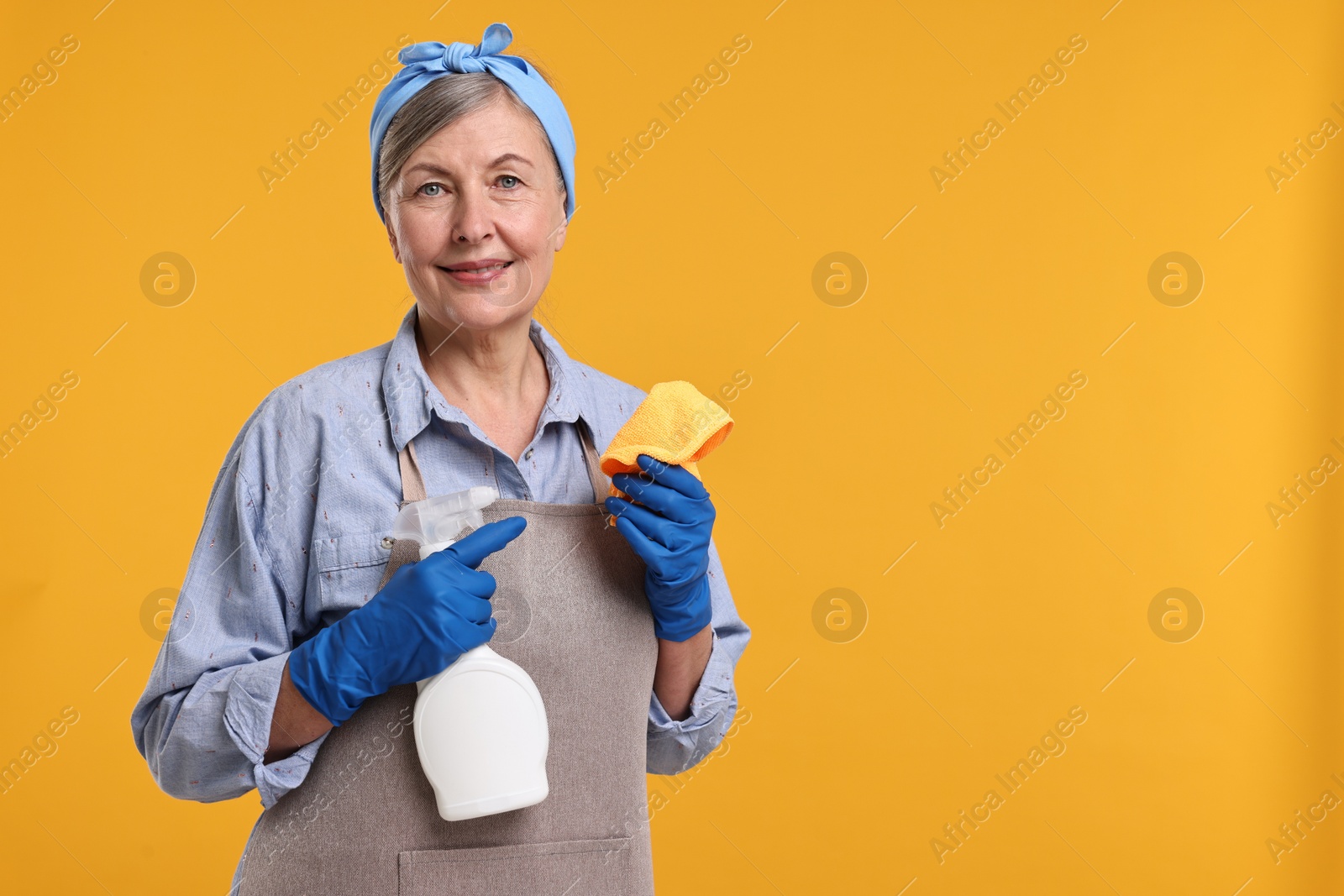 Photo of Happy housewife with spray bottle and rag on orange background, space for text