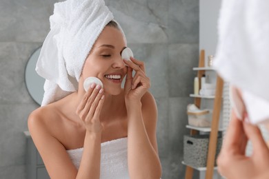 Photo of Young woman cleaning her face with cotton pads near mirror in bathroom