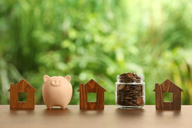 Photo of Piggy bank, jar of coins and house models on wooden table outdoors