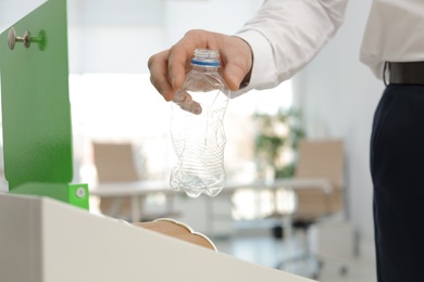 Photo of Man putting used plastic bottle into trash bin in office, closeup. Waste recycling