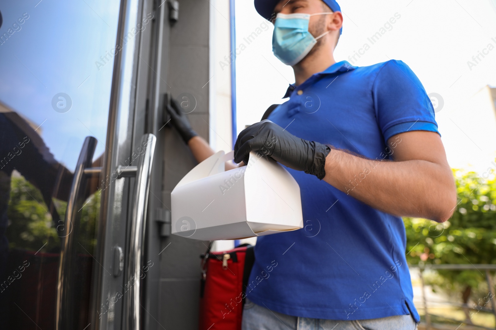Photo of Courier in protective mask and gloves with order near front door, focus on hands. Restaurant delivery service during coronavirus quarantine