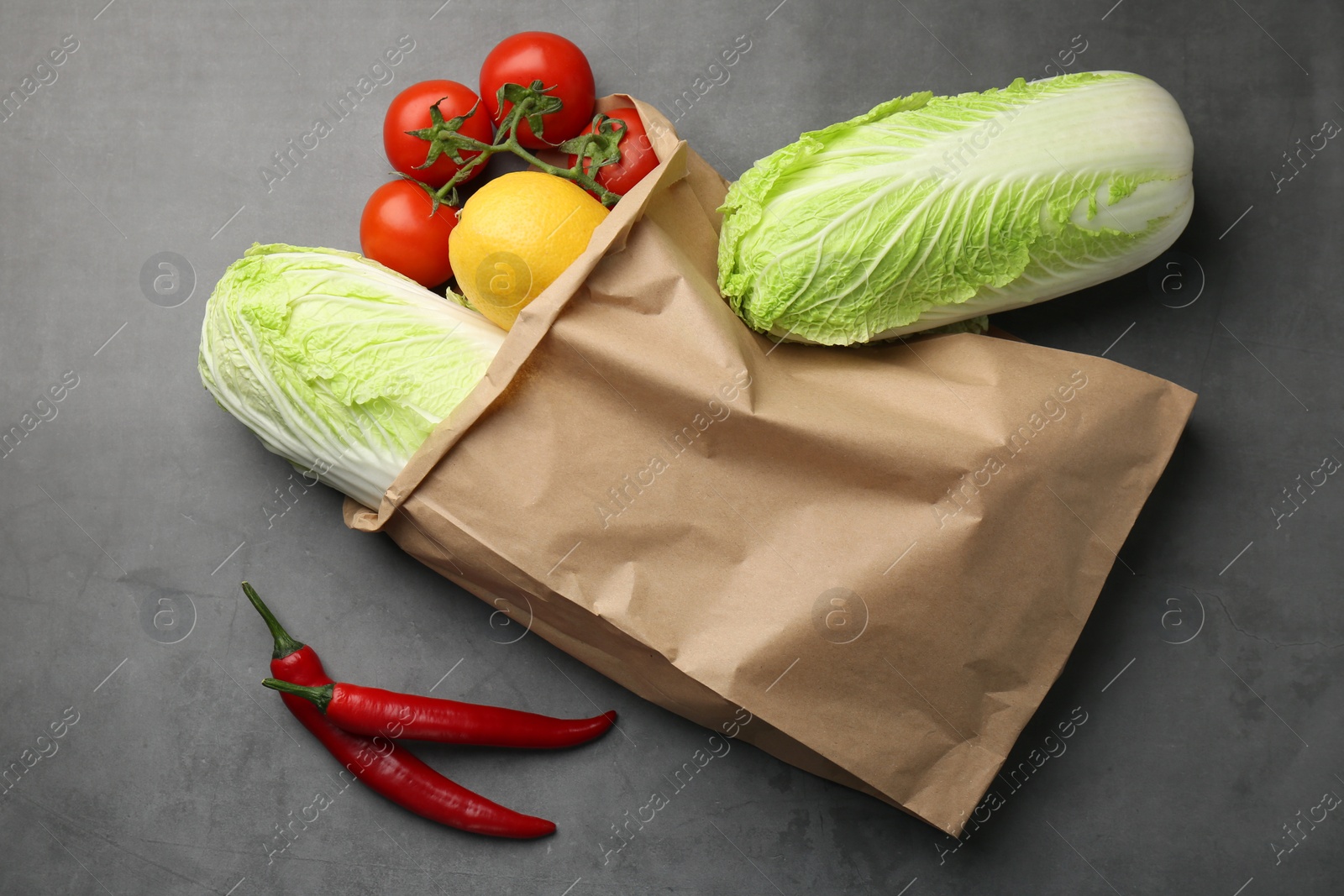 Photo of Paper bag with fresh Chinese cabbages, lemon, tomatoes and chili pepper on grey textured table, flat lay