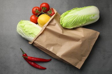 Paper bag with fresh Chinese cabbages, lemon, tomatoes and chili pepper on grey textured table, flat lay