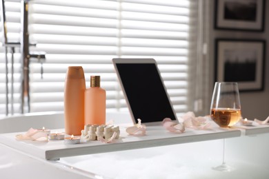 Photo of Wooden tray with tablet, wine, toiletries and flower petals on bathtub in bathroom