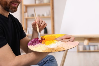 Man with brush and artist`s palette painting in studio, closeup. Using easel to hold canvas