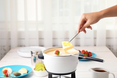 Woman dipping pineapple into pot with white chocolate fondue on table