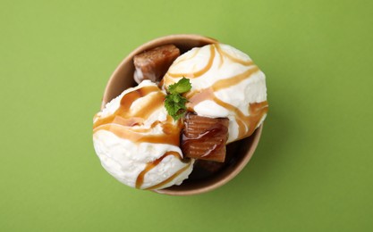 Scoops of tasty ice cream with mint leaves, caramel sauce and candies in paper cup on green background, top view