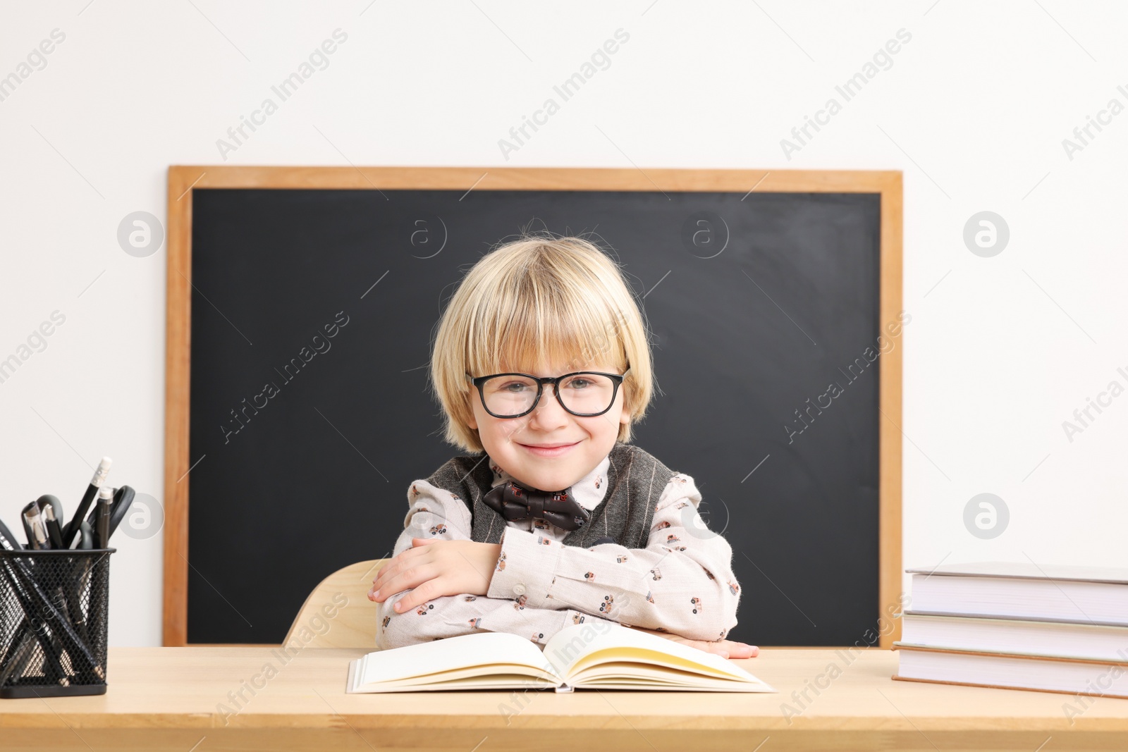 Photo of Happy little school child sitting at desk with books near chalkboard in classroom