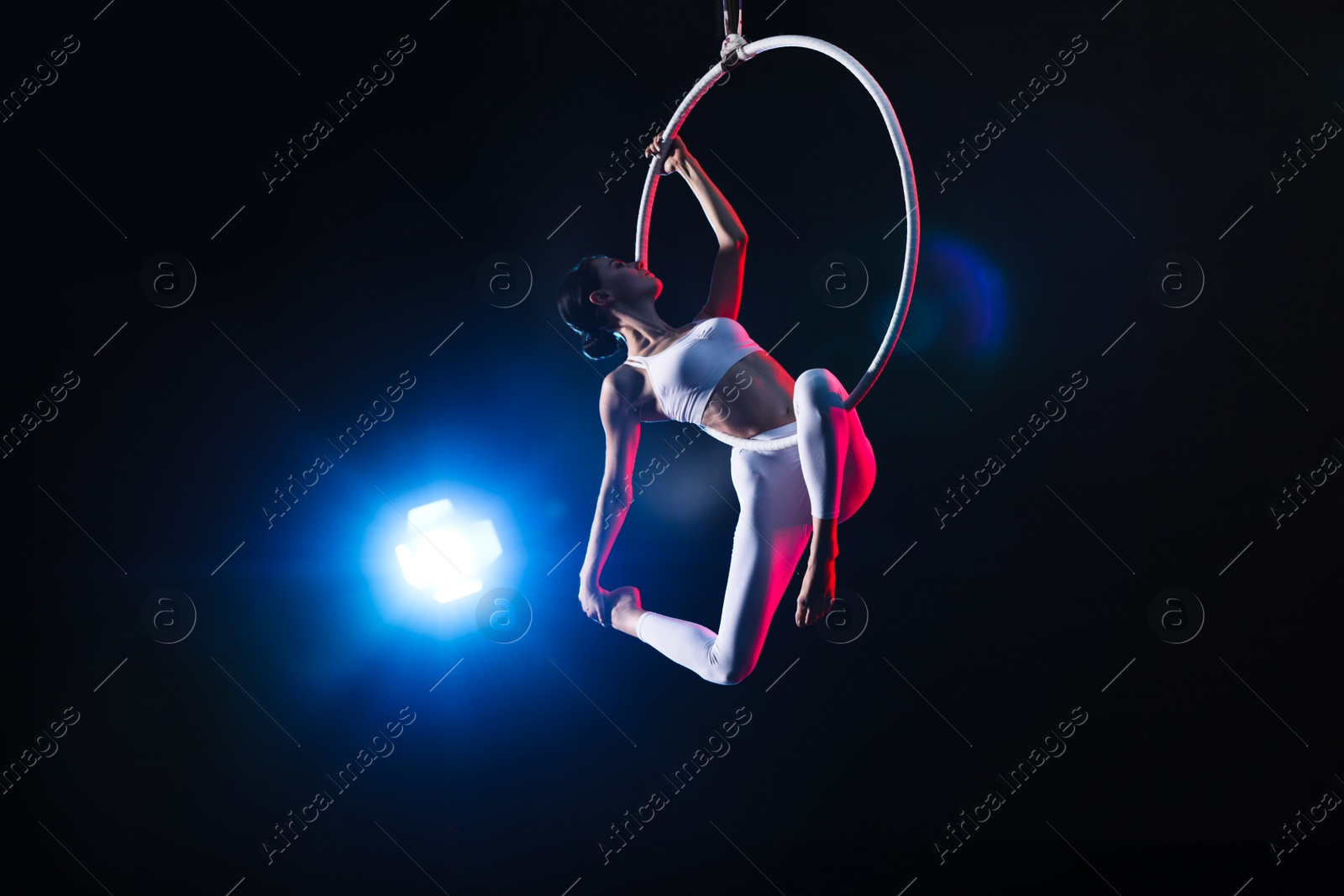 Photo of Young woman performing acrobatic element on aerial ring against dark background