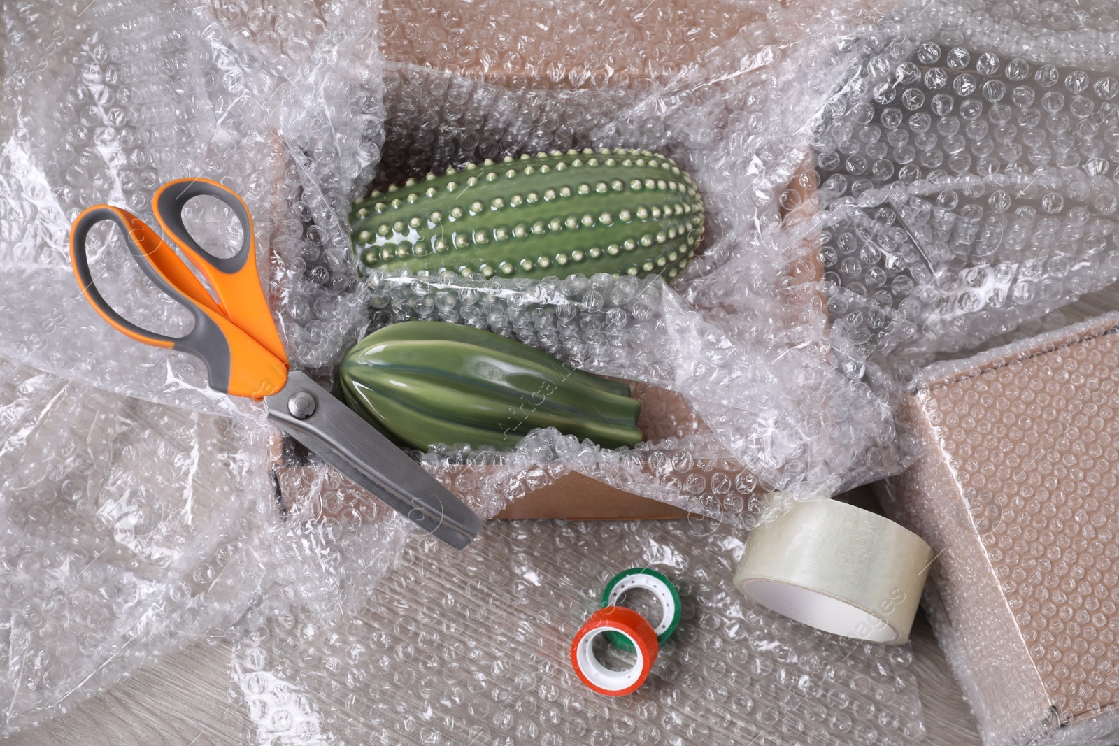 Photo of Ceramic cacti with bubble wrap in cardboard box, scissors and adhesive tapes on wooden table