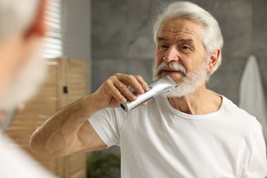 Photo of Senior man trimming beard near mirror in bathroom