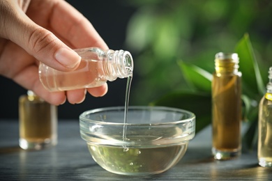 Photo of Woman pouring essential oil from glass bottle into bowl on table, closeup